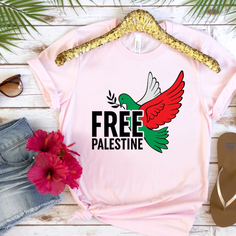 May include: Light pink t-shirt with a dove graphic in green, white, and red, holding an olive branch. The words "FREE PALESTINE" are printed in black, with the letter "E" in green. The shirt is displayed on a gold hanger.