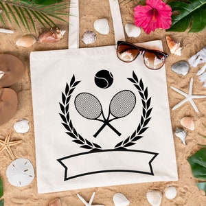 May include: White canvas tote bag with a black tennis racket and ball design. The design includes a laurel wreath and a blank banner. The bag is on a sandy surface with seashells, starfish, and tropical plants.