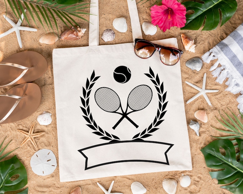 May include: White canvas tote bag with a black tennis racket and ball design. The design includes a laurel wreath and a blank banner. The bag is on a sandy surface with seashells, starfish, and tropical plants.