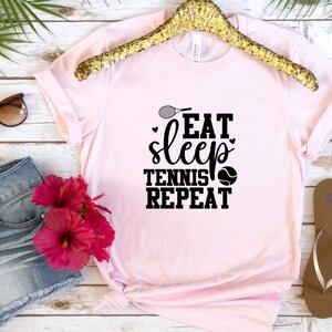 May include: Pink t-shirt with a black graphic that says "Eat Sleep Tennis Repeat" with a tennis racket and tennis ball.