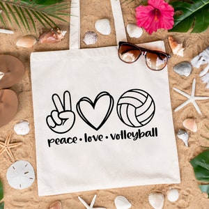 K&ouml;nnte beinhalten: Wei&szlig;e Canvas-Tragetasche mit schwarzem Text, der "peace. love. volleyball" lautet. Die Tasche zeigt ein Peace-Zeichen, ein Herz und eine Volleyball-Grafik.