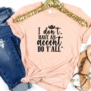 May include: A peach-colored t-shirt with the text "I don't have an accent do y'all" in black. The text is in a cursive font and includes a small cowboy hat graphic above the word "don't".