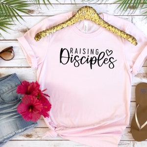May include: Pink t-shirt with black text that reads "Raising Disciples" with a small heart symbol.
