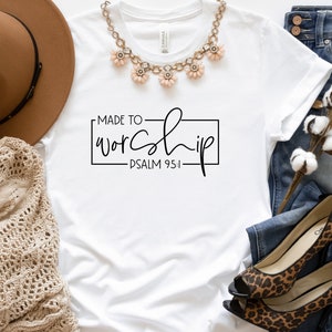 May include: White t-shirt with the text "MADE TO WORSHIP - PSALM 95:1" in black lettering. The shirt is laid flat on a white surface with a brown hat, a necklace, a pair of jeans, and a cotton plant.