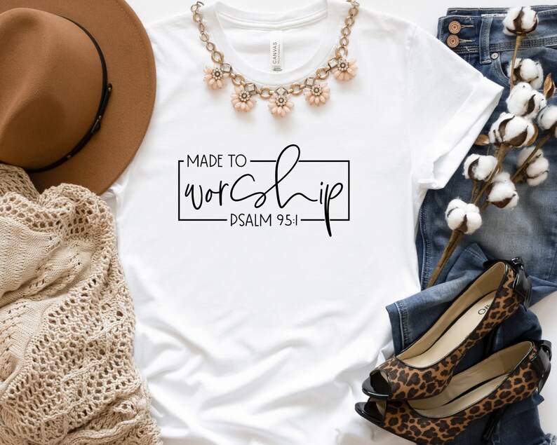 May include: White t-shirt with the text "MADE TO WORSHIP - PSALM 95:1" in black lettering. The shirt is laid flat on a white surface with a brown hat, a necklace, a pair of jeans, and a cotton plant.