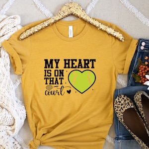 May include: A mustard yellow t-shirt with the text "My heart is on that court" and a green softball in a heart shape.