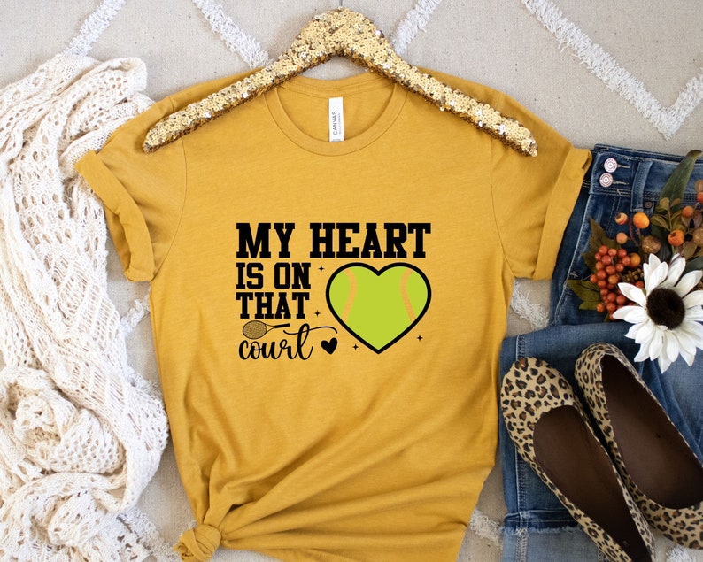 May include: A mustard yellow t-shirt with the text "My heart is on that court" and a green softball in a heart shape.