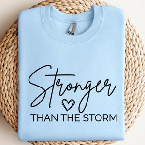 May include: Light blue sweatshirt with the text "Stronger than the storm" in black cursive lettering with a small heart symbol.