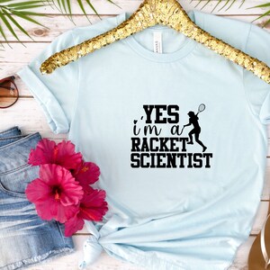 May include: A light blue t-shirt with a black graphic that says "YES i'm a RACKET SCIENTIST" and a silhouette of a person playing tennis.