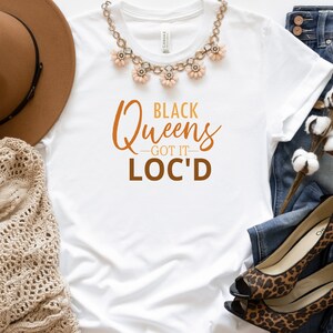 May include: White t-shirt with the text "Black Queens - Got It - Loc'd" in brown and orange lettering. The shirt is lying on a white surface with a brown hat, a crocheted shawl, a pair of jeans, and a pair of leopard print heels.