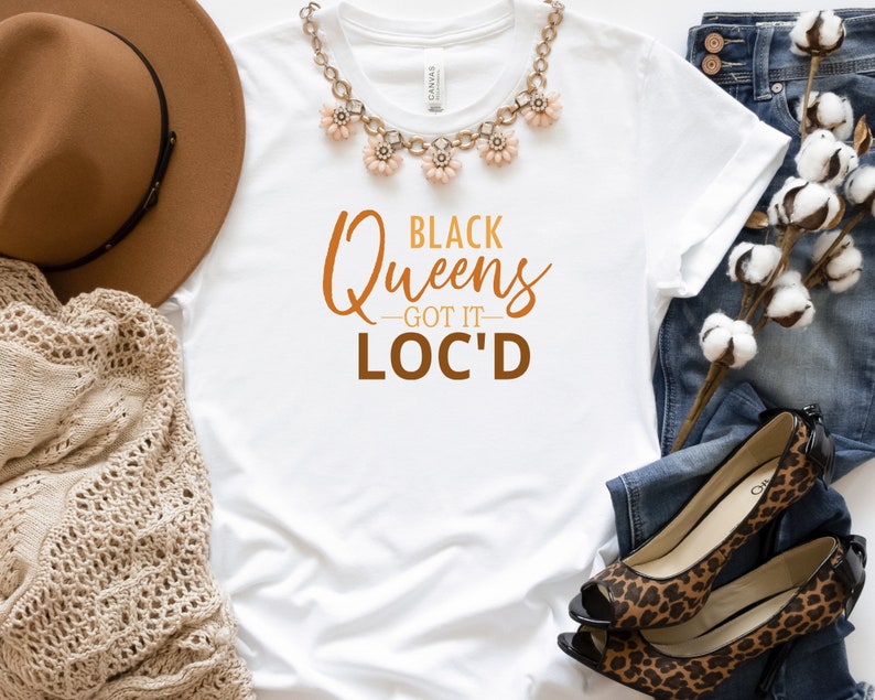 May include: White t-shirt with the text "Black Queens - Got It - Loc'd" in brown and orange lettering. The shirt is lying on a white surface with a brown hat, a crocheted shawl, a pair of jeans, and a pair of leopard print heels.
