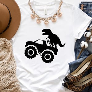 May include: White t-shirt featuring a black silhouette of a monster truck with a dinosaur on top.