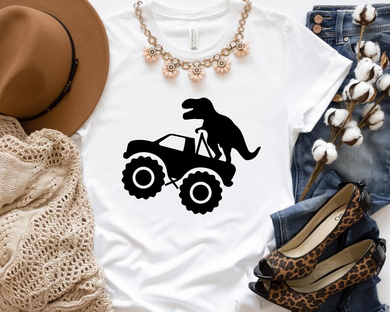 May include: White t-shirt featuring a black silhouette of a monster truck with a dinosaur on top.