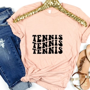 May include: A light pink t-shirt with a black graphic that says "TENNIS" three times in a retro font.