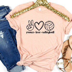K&ouml;nnte beinhalten: Ein pfirsichfarbenes T-Shirt mit einem Peace-Zeichen, einem Herzen und einem Volleyball-Motiv. Der Text "peace. love. volleyball" ist unter dem Motiv aufgedruckt.