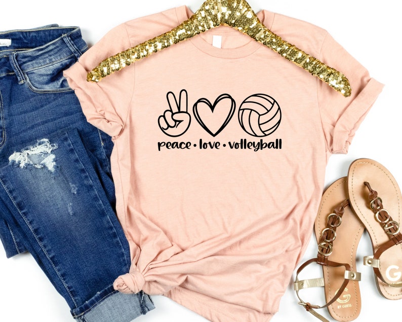 K&ouml;nnte beinhalten: Ein pfirsichfarbenes T-Shirt mit einem Peace-Zeichen, einem Herzen und einem Volleyball-Motiv. Der Text "peace. love. volleyball" ist unter dem Motiv aufgedruckt.