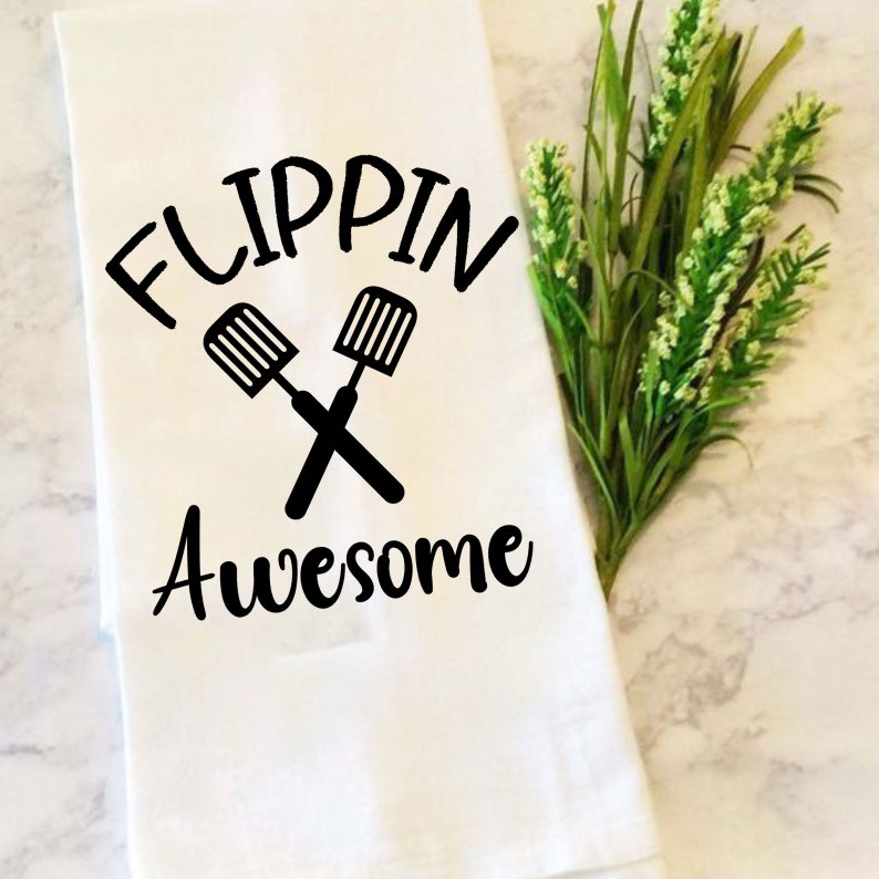 K&ouml;nnte beinhalten: Ein wei&szlig;es Geschirrtuch mit schwarzem Text, der "Flippin' Awesome" lautet, mit zwei gekreuzten Spateln &uuml;ber dem Text.