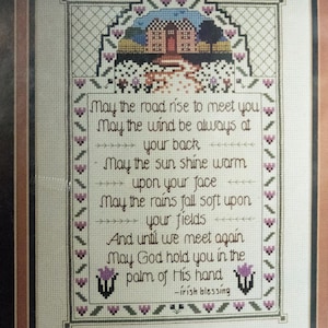 May include: Cross-stitch artwork featuring an Irish blessing. The framed piece includes a detailed border and a central text with a house and landscape scene at the top. The text reads "May the road rise to meet you..." in a decorative font.