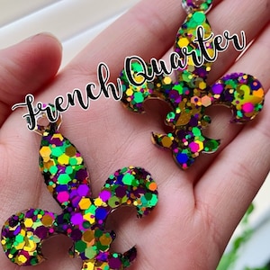 May include: Two gold glitter fleur-de-lis charms with purple, green, and pink glitter accents. The charms are on a hand and the text "French Quarter" is written across the image.