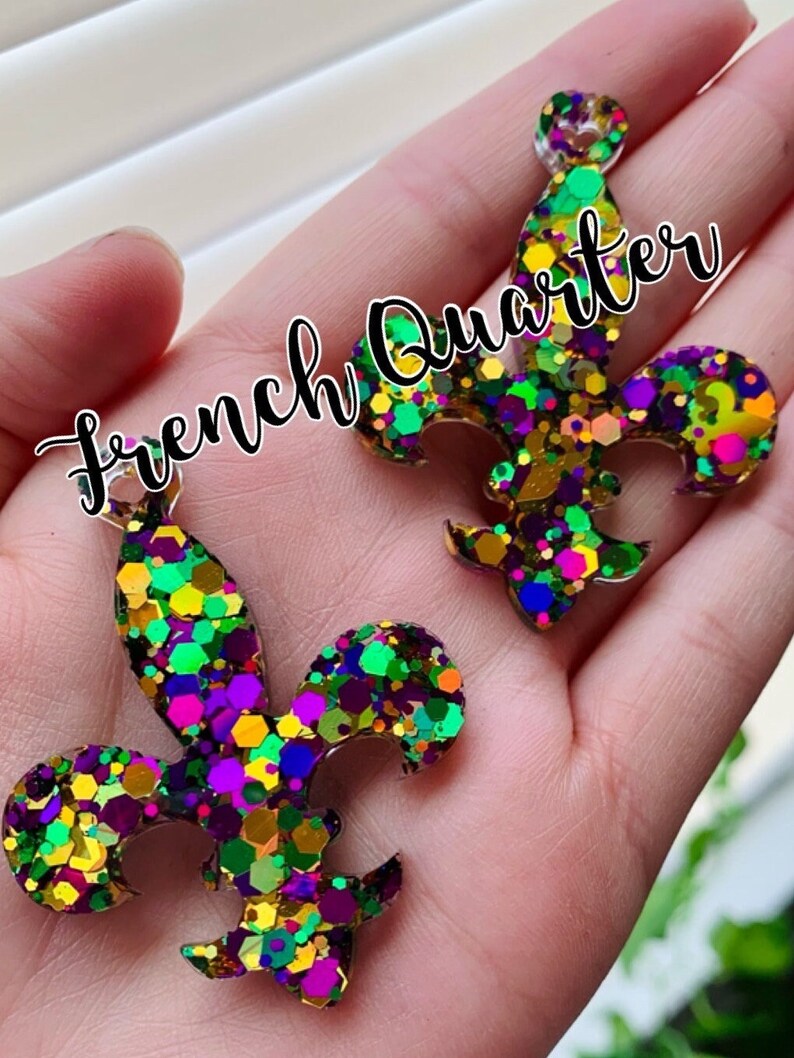 May include: Two gold glitter fleur-de-lis charms with purple, green, and pink glitter accents. The charms are on a hand and the text "French Quarter" is written across the image.