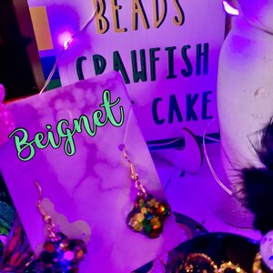 May include: A pair of gold earrings with a multi-coloured, glittery star charm. The earrings are displayed on a purple card with the word "Bignet" written in green. The card is in front of a sign that says "Beads Crawfish Cake".
