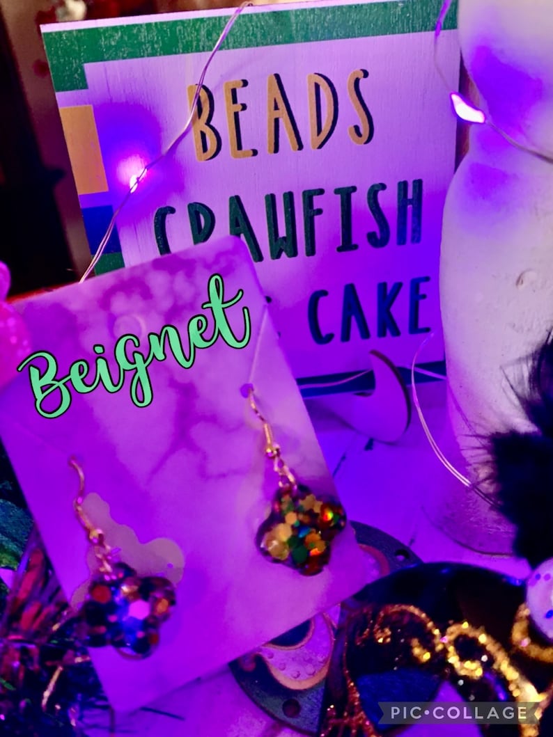 May include: A pair of gold earrings with a multi-coloured, glittery star charm. The earrings are displayed on a purple card with the word "Bignet" written in green. The card is in front of a sign that says "Beads Crawfish Cake".