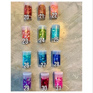 May include: A collection of 11 small jars filled with colorful chunky glitter. The jars are labeled with numbers 20 through 31.