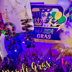 May include: A pair of gold earrings with a Mardi Gras theme. The earrings are shaped like the letters "G" and "R" and are covered in colourful glitter. The earrings are hanging from a white marble background with the words "Mardi Gras" in purple and gold letters.