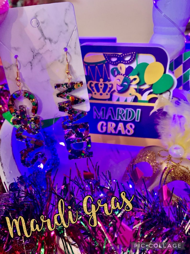 May include: A pair of gold earrings with a Mardi Gras theme. The earrings are shaped like the letters "G" and "R" and are covered in colourful glitter. The earrings are hanging from a white marble background with the words "Mardi Gras" in purple and gold letters.