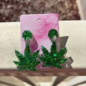 May include: A pair of green glitter dangle earrings shaped like marijuana leaves. The earrings are on a pink background.