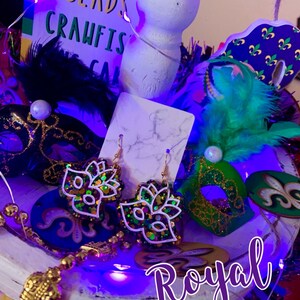 May include: A pair of Mardi Gras themed earrings with gold hooks and a green, purple, and gold glitter mask design. The earrings are displayed on a white surface with other Mardi Gras decorations, including a purple, green, and gold mask, a feather boa, and a sign that says "Beads Crawfish Cake".