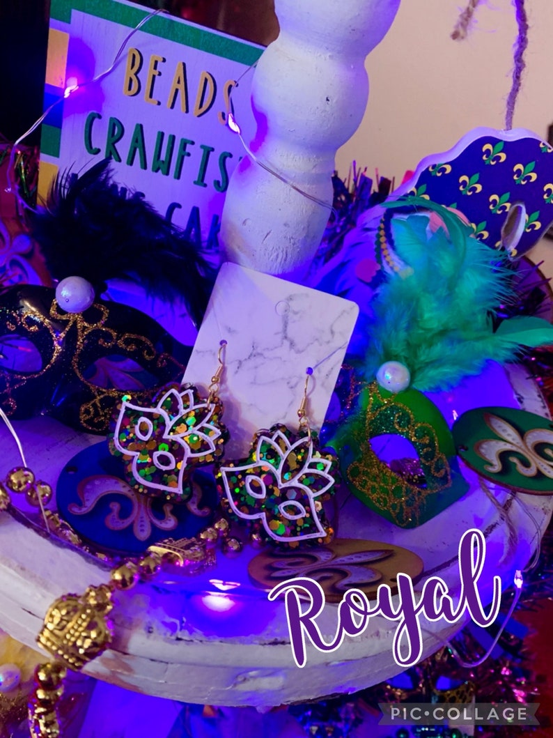 May include: A pair of Mardi Gras themed earrings with gold hooks and a green, purple, and gold glitter mask design. The earrings are displayed on a white surface with other Mardi Gras decorations, including a purple, green, and gold mask, a feather boa, and a sign that says "Beads Crawfish Cake".