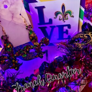 May include: Purple, green and gold Mardi Gras themed earrings with a fleur-de-lis design. The earrings are displayed on a white background with a sign that says "LOVE" and a fleur-de-lis. The sign is surrounded by purple, green and gold Mardi Gras decorations.