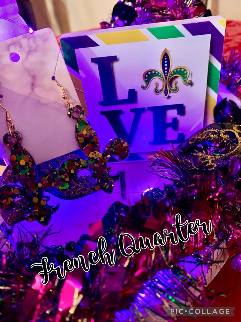 May include: Purple, green and gold Mardi Gras themed earrings with a fleur-de-lis design. The earrings are displayed on a white background with a sign that says "LOVE" and a fleur-de-lis. The sign is surrounded by purple, green and gold Mardi Gras decorations.