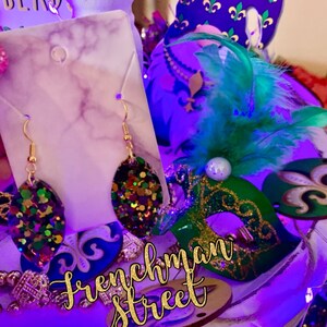 May include: Gold earrings with a green, purple and gold glitter oval design. The earrings are displayed on a white marble background with the text "Frenchman Street" in gold glitter letters.