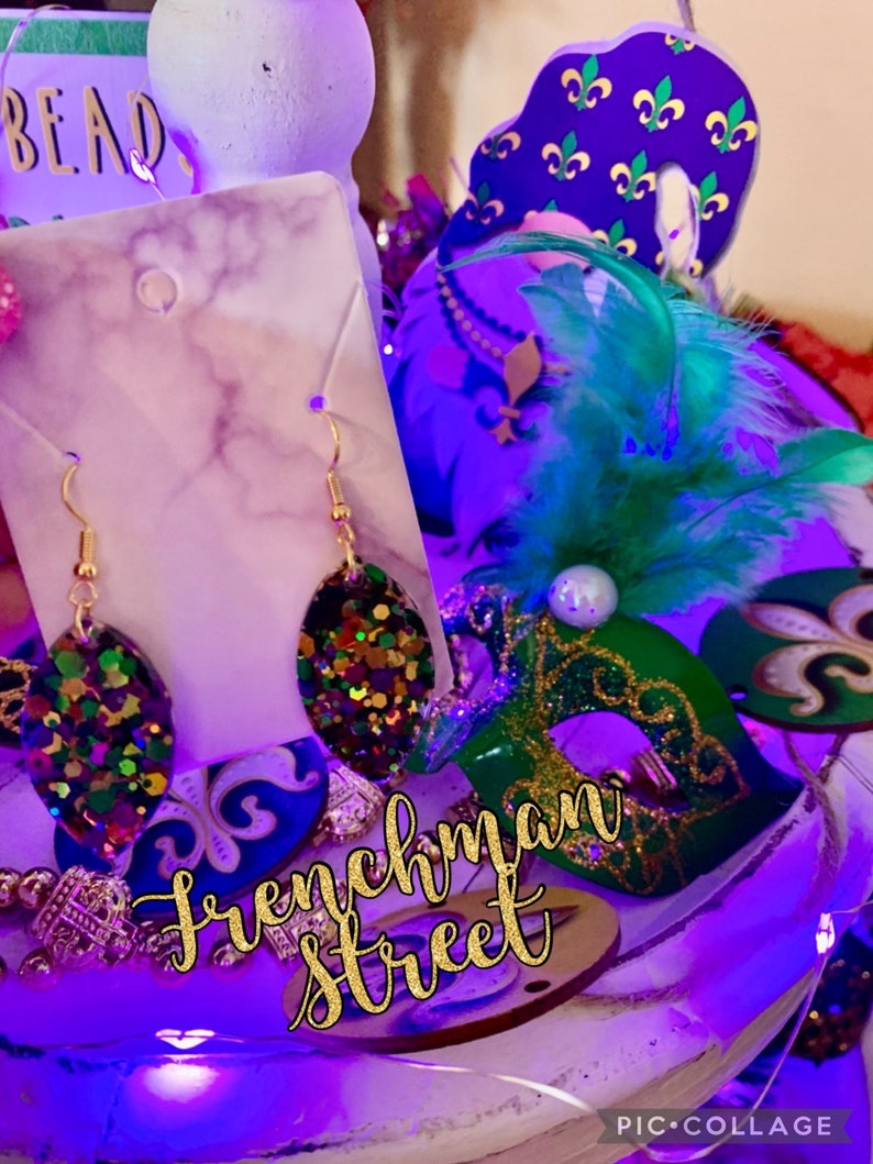 May include: Gold earrings with a green, purple and gold glitter oval design. The earrings are displayed on a white marble background with the text "Frenchman Street" in gold glitter letters.