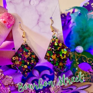 May include: A pair of gold dangle earrings with a diamond-shaped resin charm. The charms are filled with colourful glitter, including green, purple, gold, and red. The earrings are labelled "Bourbon Street".
