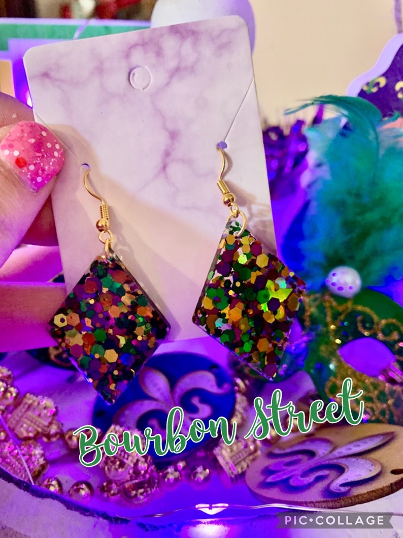 May include: A pair of gold dangle earrings with a diamond-shaped resin charm. The charms are filled with colourful glitter, including green, purple, gold, and red. The earrings are labelled "Bourbon Street".
