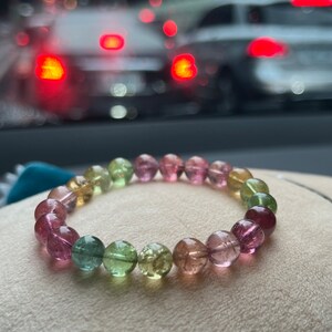 May include: A multi-colored gemstone bracelet with pink, green, and yellow beads. The bracelet is made of round, smooth beads and is lying on a light brown surface.