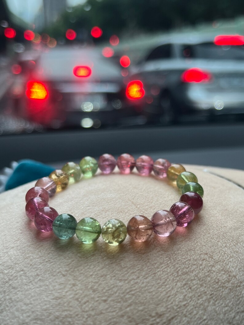May include: A multi-colored gemstone bracelet with pink, green, and yellow beads. The bracelet is made of round, smooth beads and is lying on a light brown surface.