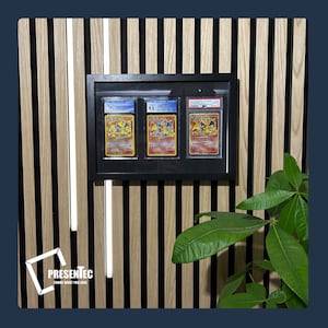 May include: A black frame with three Pokemon cards inside. The cards are graded and have a white background with colorful illustrations. The frame is mounted on a wall with wood paneling.