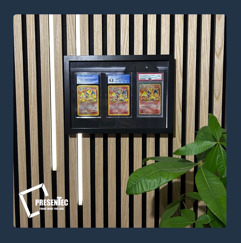 May include: A black frame with three Pokemon cards inside. The cards are graded and have a white background with colorful illustrations. The frame is mounted on a wall with wood paneling.