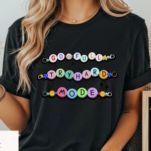 May include: Black t-shirt with the words "GO FULL", "TRY HARD", and "MODE" in colorful, circular letters. A small chalkboard displays the text "I believe in you!" with a star and lightbulb graphic.