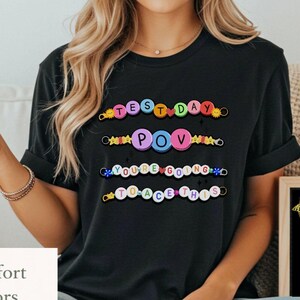 May include: Black t-shirt with the phrases "TEST DAY POV YOU'RE GOING TO ACE THIS" in colorful, beaded letters. A small chalkboard reads "I believe in you!" with a star and lightbulb graphic. The shirt also has the words "Comfort Colors" on it.