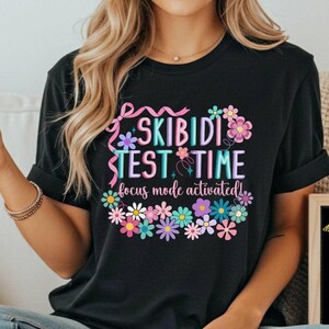 May include: Black t-shirt with the text "SKIBIDI TEST TIME focus mode activated!" in a floral design. A small chalkboard displays the message "I believe in you!" with a star and lightbulb graphic.