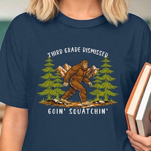 May include: Navy blue t-shirt with a graphic of Bigfoot walking in front of pine trees and mountains. Text reads "Third Grade Dismissed, Goin' Squatchin'". The person is holding two books.