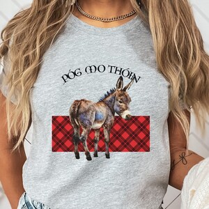 May include: A gray t-shirt featuring a donkey illustration with the text "Póg mo thóin" above a red plaid design. The donkey is brown, blue, and white. The shirt is a crew neck.