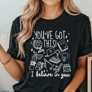 Puede incluir: Camiseta negra con el texto "YOU'VE GOT THIS! I believe in you" e ilustraciones escolares en blanco. Una peque&ntilde;a pizarra muestra "I believe in you!" con una estrella y una bombilla. La frase "10 designs available" est&aacute; en la parte inferior.