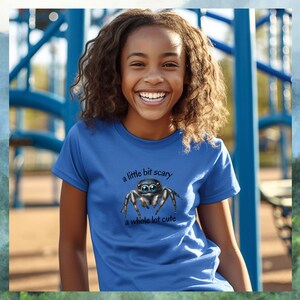May include: A blue t-shirt with the text "a little bit scary a whole lot cute" and a graphic of a jumping spider. The person wearing the shirt is smiling. The background is a playground.