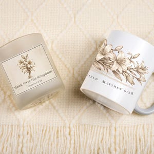 May include: A white ceramic mug and a frosted glass candle holder with floral designs. The mug has a gray handle and the text "Matthew 6:28". The candle holder has the text "Seek First His Kingdom Matthew 6:33".